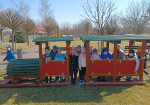 Grupa dzieci stoi w pociągu na placu zabaw, chłopiec trzyma maskotkę krasnala Hałabały.